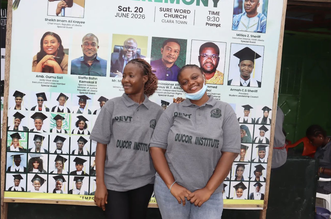 Two DEVT graduates standing together in front of the graduation ceremony banner at the institute's graduation