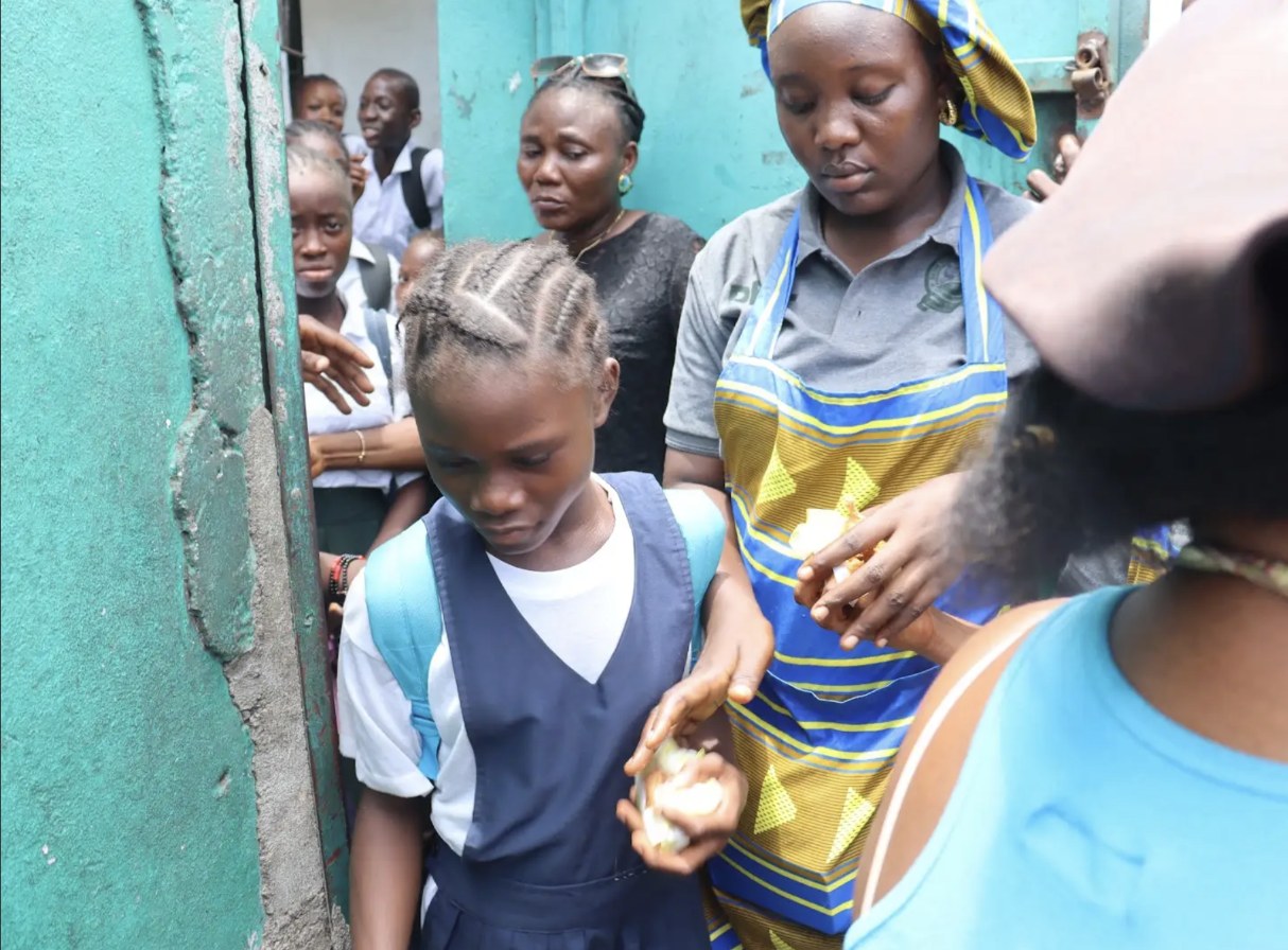 A DEVT volunteer in a branded apron handing a free hot meal to a public school student in uniform during Ducor Service Month in District #14